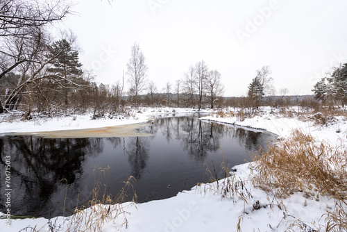 Dark river against a background of white snow. Cloudy winter evening. Dry old grass sticks out from under the snow © Sergei