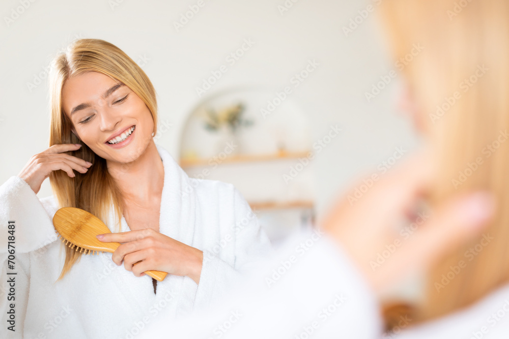 Pretty blonde woman untangles and brushing hair standing in bathroom