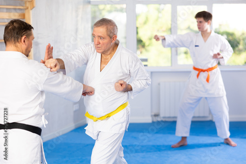 Focused experienced elderly karate fighter engaging kumite with male rival, demonstrating martial art techniques in training room