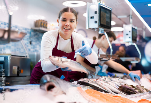 Positive young saleswoman demonstrating seabass to client in fish store with large stock
