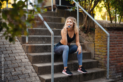 Smilling fitness woman in sportswear eating fitness bar while sitting on stairs outdoors. Blonde sporty girl resting after workout. Healthy and sport concept