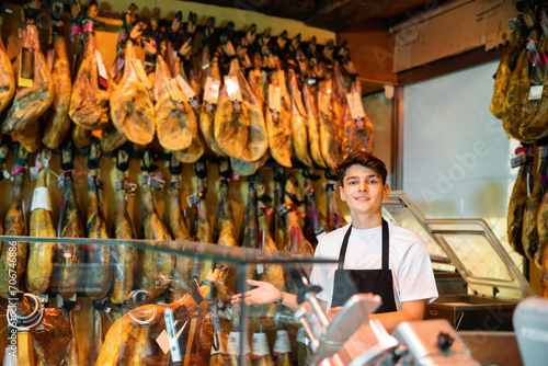 Caucasian young man in uniform standing at jamoneria counter, gesturing and looking at camera.