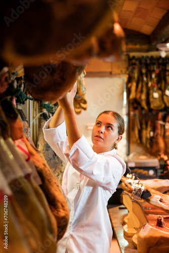 Portrait of positive young salesgirl of small local butcher shop specializing in sale of jerky Iberian jamon working behind counter