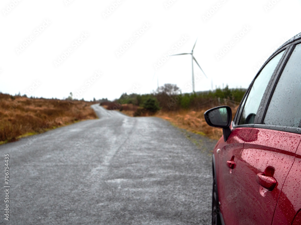 Red car parked off small rough country road, Wind power generators in a forest against white sky background. Travel to nature to see green energy technology concept.