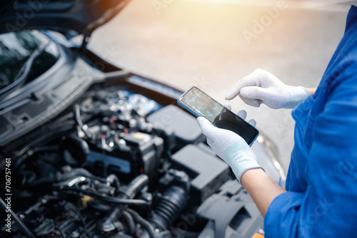 Mechanic examining car engine with mobile phone at auto repair shop. Close-up shot of male technician taking a break and texting on smartphone. Horizontal photo with workshop background.