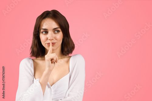 A young, confident woman in a white blouse holds a finger to her lips in a hushing gesture