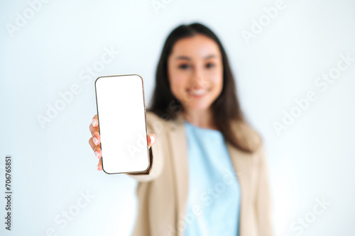 Defocused multiracial woman, manager, stands on isolated white background, showing smartphone with white blank mock-up screen for advertising or presentation, smiling