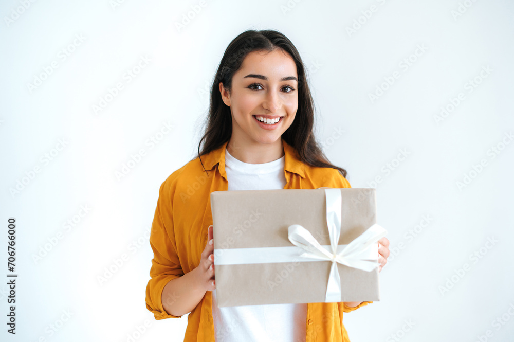Gift box. Happy pretty brunette indian or arabian woman in casual orange shirt, stands on isolated white background, holding big gift box, looking at camera and smiles friendly Gift box. Happy pretty brunette indian or arabian woman in casual orange shirt, stands on isolated white background, holding big gift box, looking at camera and smiles friendly