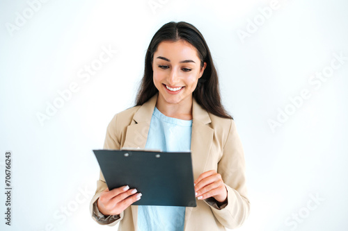 Happy beautiful brunette indian or arabian woman, hiring manager, financial consultant stands on isolated white background, holding folder with documents or resume, looking at it, smiling friendly