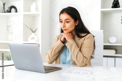 Serious concentrated young adult indian or arabian business lady, executive, company employee, lawyer, in formal wear, sitting in modern office, focused looks at laptop screen, studying information