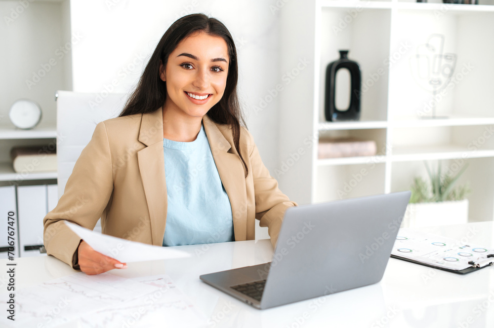 Cute attractive young adult indian or arabian business woman entrepreneur, financial expert of company, wearing suit, sitting in the office, holding financial documents, looking at camera and smiles Cute attractive young adult indian or arabian business woman entrepreneur, financial expert of company, wearing suit, sitting in the office, holding financial documents, looking at camera and smiles