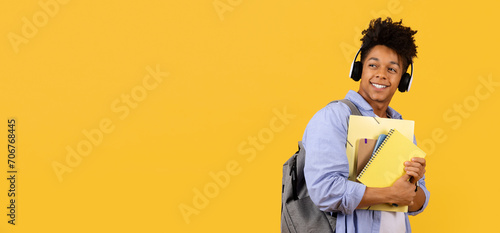 Smiling black male student with headphones and notebooks on yellow