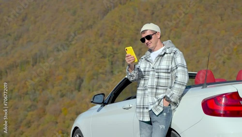 Successful happy guy watches at smartphone stands near whate car in mountains. photo