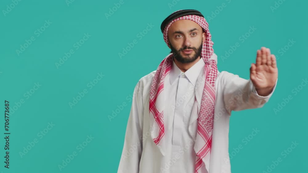 Middle eastern model doing stop symbol with palm raised, expressing negativity and rejection on camera. Young adult showcasing refusal, being displeased with something in studio. Middle eastern model doing stop symbol with palm raised, expressing negativity and rejection on camera. Young adult showcasing refusal, being displeased with something in studio.