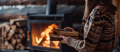 Stylish woman in knitted sweater putting firewood to modern black fireplace with warm fire Cozy warm moments at cold season Hipster female relaxing in comfortable cabin in mountains