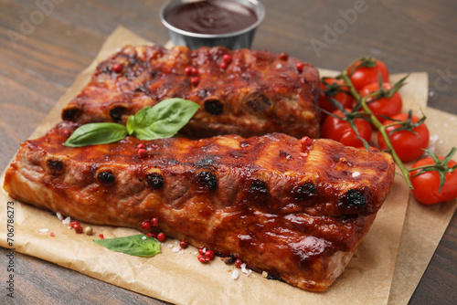 Tasty roasted pork ribs served with sauce, basil and tomatoes on wooden table, closeup