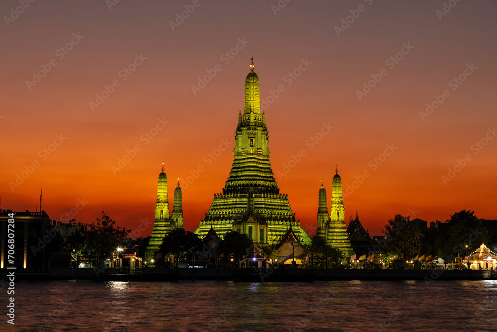 Fototapeta premium Wat Arun (Temple of the dawn) in Bangkok at dusk. Temple and prangs bathed in yellow light. Deepening orange sky in background. Chao Phraya river in foreground. 