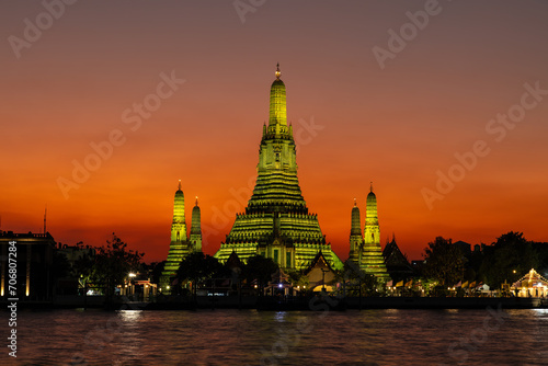 Wat Arun (Temple of the dawn) in Bangkok at dusk. Temple and prangs bathed in yellow light. Deepening orange sky in background. Chao Phraya river in foreground. 
