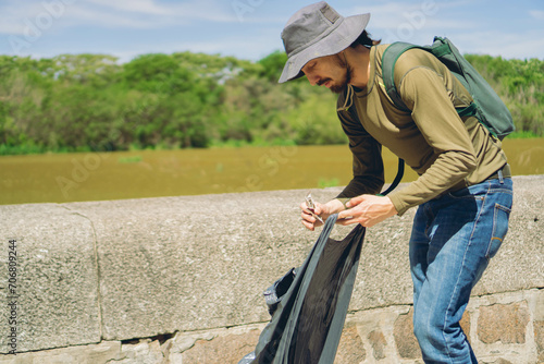 Young Latin man picks up garbage from an ecological reserve on a sunny day, recycling concept