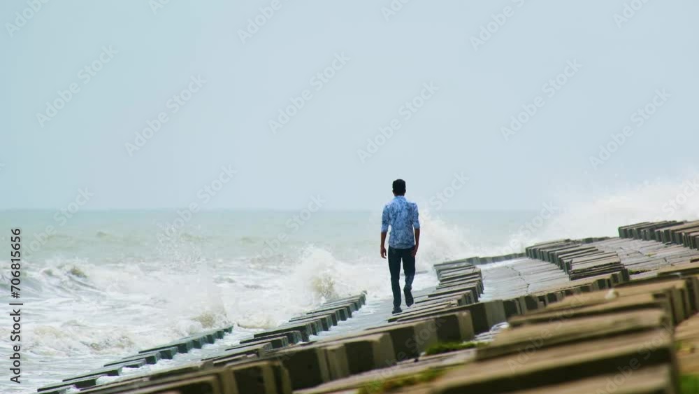 Man appearing to be depressed and lonely walking at sea shore in slow motion against the crushing waves on a concrete breakwater slope.