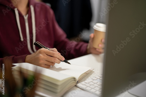 Cropped image of a man sitting in front of a computer, taking notes in a book, working in a room. photo
