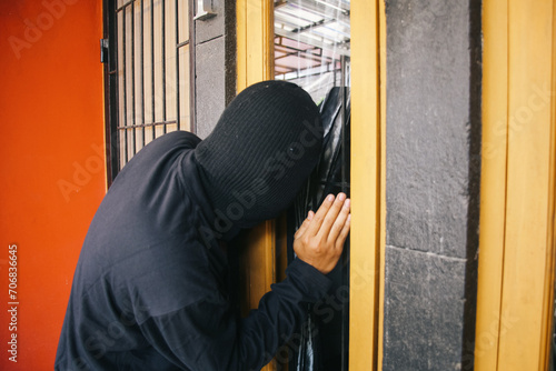 Burglar wearing a balaclava peeking and looking through the house window. Criminal and security concept.
