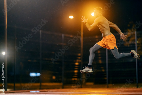 An urban shirtless muscular runner is jumping and running on cold rainy weather at night.