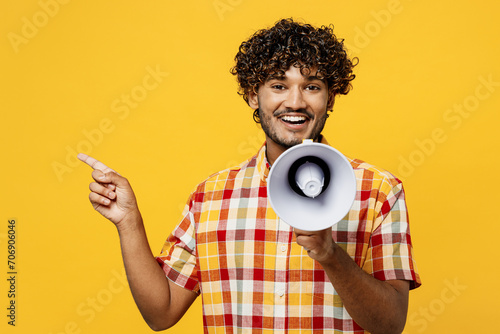 Young Indian man he wears shirt casual clothes hold in hand megaphone scream announces discounts sale Hurry up point aside isolated on plain yellow color background studio portrait. Lifestyle concept.