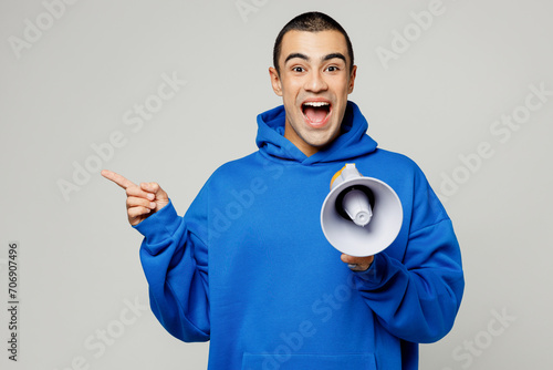 Young surprised middle eastern man wears blue hoody casual clothes old in hand megaphone scream announces discounts sale Hurry up point finger aside on area isolated on plain solid white background.