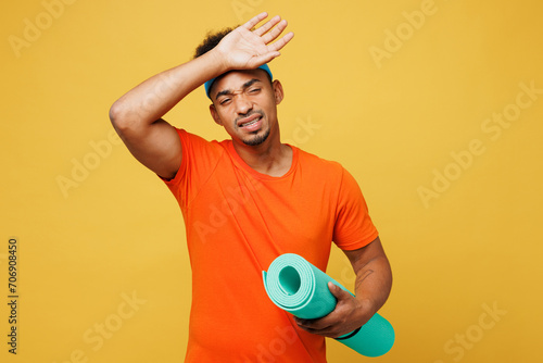 Young tired sad fitness trainer instructor sporty man sportsman wear orange t-shirt put hand on head hold yoga mat train in home gym isolated on plain yellow background. Workout sport fit abs concept. photo