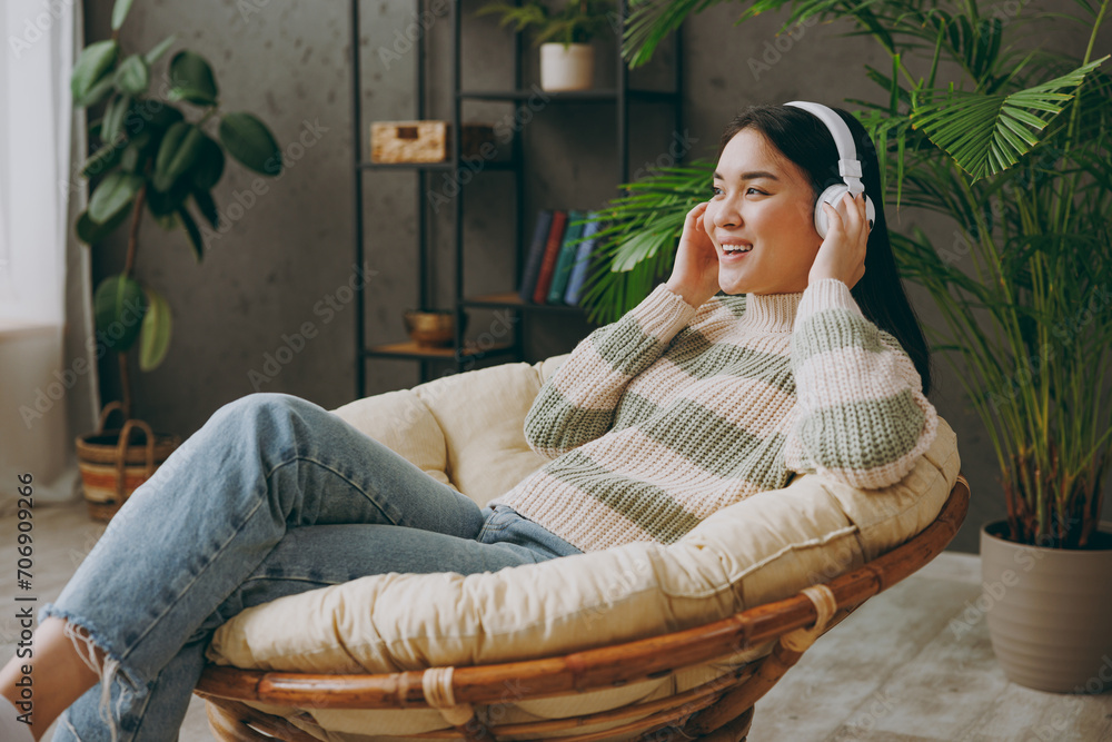 Obraz premium Sideways young woman she wear casual clothes listen to music in headphones sits on armchair stays at home hotel flat rest relax spend free spare time in grey living room indoor. People lounge concept.