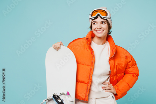 Young smiling woman wear warm padded windbreaker jacket hat ski goggles mask hold snowboard look aside on area travel rest spend weekend winter season in mountains isolated on plain blue background.