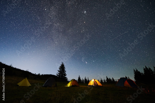 Night camping in mountains under starry sky. Tourist tents in campsite under beautiful sky full of stars with Milky way above forest. Concept of tourism and traveling.