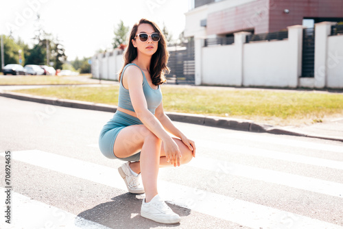 Young beautiful smiling female in trendy summer blue cycling shorts and tank top clothes. Carefree woman posing in street. Positive model having fun. Cheerful and happy. Sits on asphalt