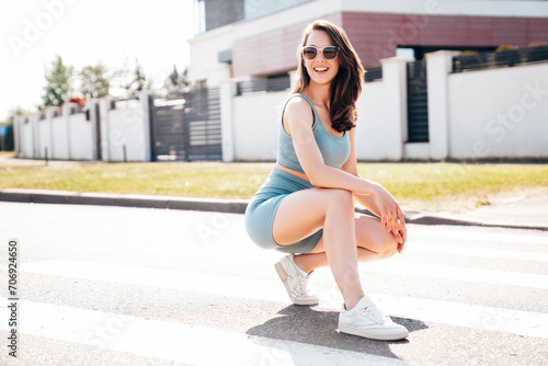 Young beautiful smiling female in trendy summer blue cycling shorts and tank top clothes. Carefree woman posing in street. Positive model having fun. Cheerful and happy. Sits on asphalt