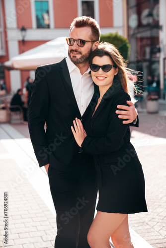 Beautiful fashion woman and her handsome elegant boyfriend in black suit. Sexy brunette model in jacket. Fashionable confident couple posing in street. Brutal man and female outdoors. In sunglasses