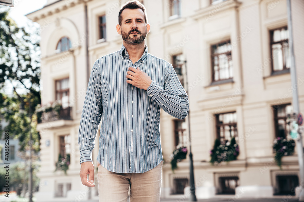 Fototapeta premium Portrait of handsome confident stylish hipster lambersexual model. Sexy modern man dressed in blue shirt and trousers. Fashion male posing on street background in Europe city at sunset