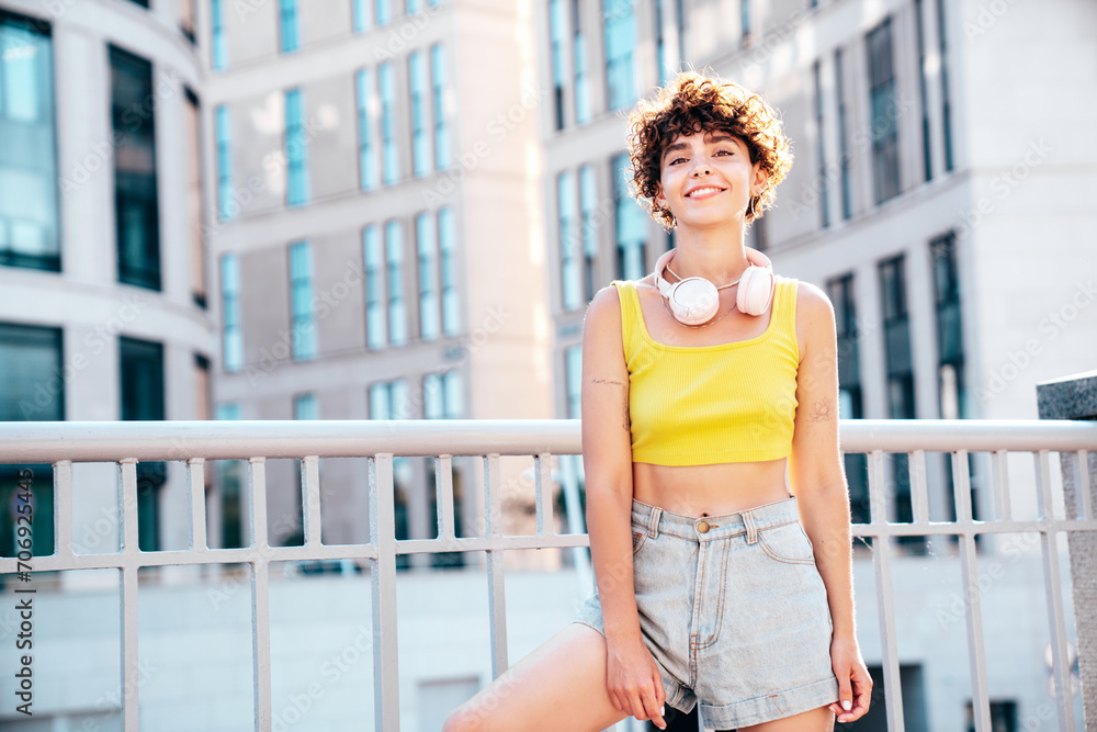 Young beautiful smiling hipster woman in trendy summer clothes. Carefree woman with curls hairstyle, posing in the street at sunny day. Positive model outdoors. Listens music at her earphones