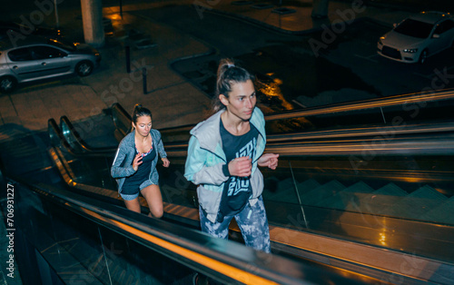 Two young women friends training running up escalator in the city at night. Selective focus on woman in background.