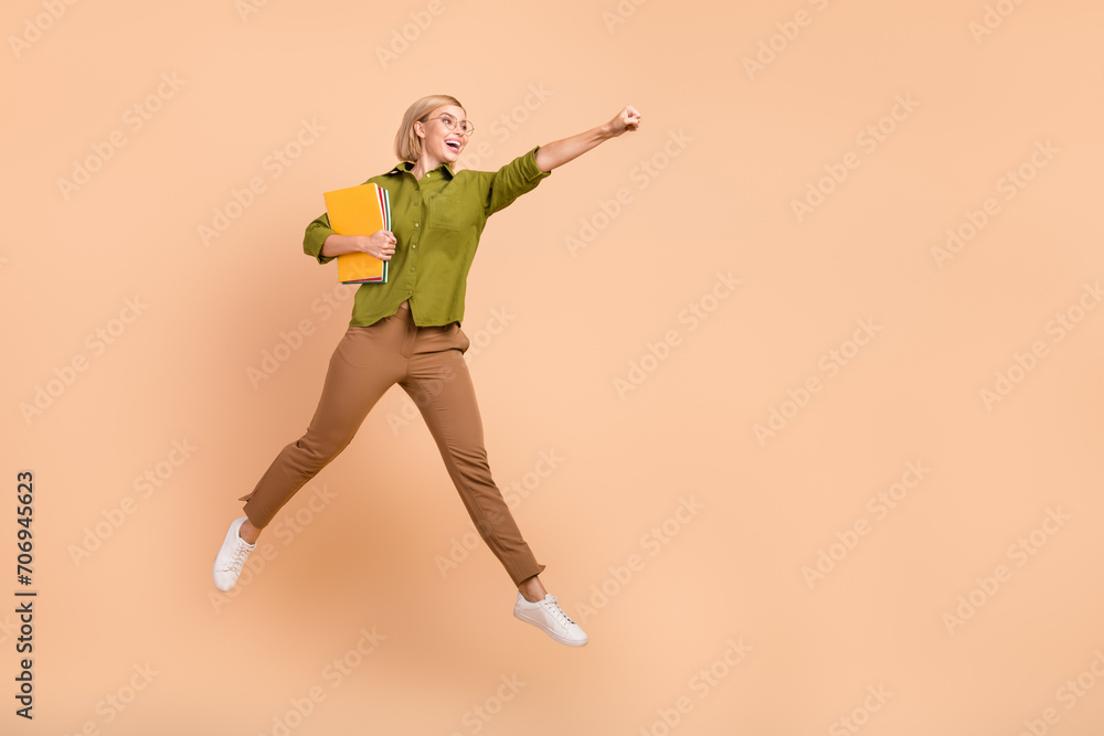Full size photo of nice girl wear khaki shirt flying hold copybooks clenching fist look empty space isolated on pastel color background