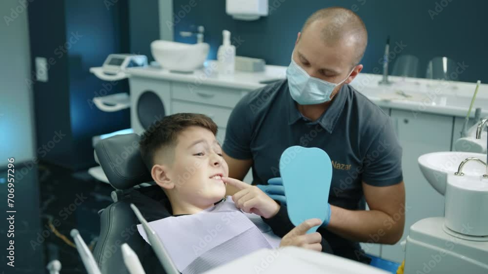 Young male dental expert giving recommmendations to young patient sitting in dentist chair with napkin on chest, holding hand mirror, showing sick tooth. High quality 4k footage Young male dental expert giving recommmendations to young patient sitting in dentist chair with napkin on chest, holding hand mirror, showing sick tooth. High quality 4k footage