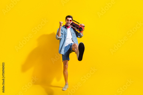 Full size photo of overjoyed ecstatic guy wear jeans jacket shorts in sunglass hold boombox show shoes isolated on yellow color background photo