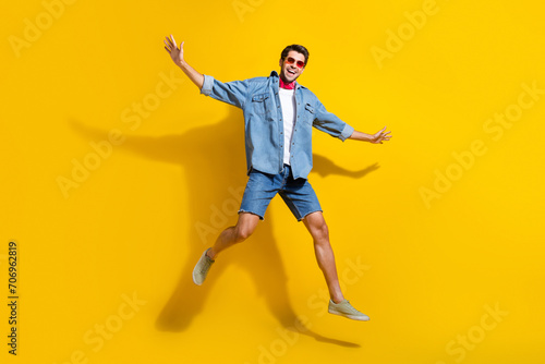 Full size photo of good mood guy wear jeans jacket in sunglass jumping hold arms like wings isolated on vibrant yellow color background