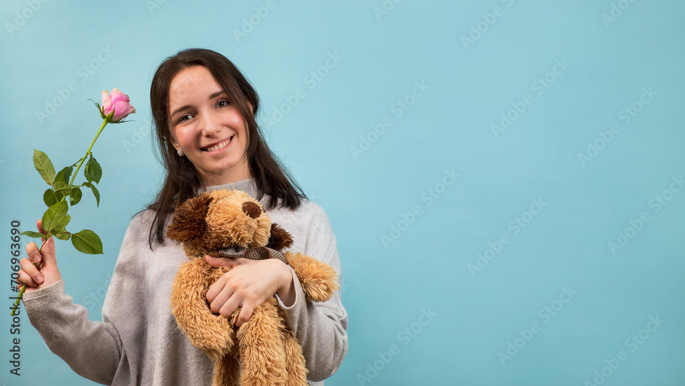 A young smiling woman holding a stuffed toy and a pink rose. Blue cardboard background with copy space for text. A young smiling woman holding a stuffed toy and a pink rose. Blue cardboard background with copy space for text.