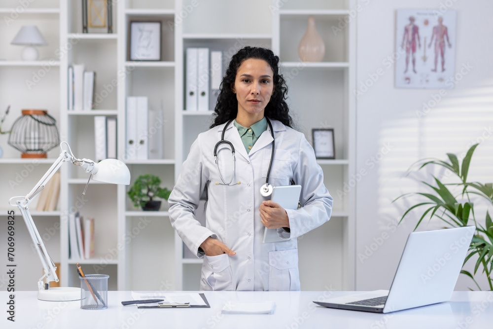 Portrait of serious confident female doctor inside medical office of hospital, Hispanic woman in white medical coat standing near desk, holding tablet computer, looking at camera. Portrait of serious confident female doctor inside medical office of hospital, Hispanic woman in white medical coat standing near desk, holding tablet computer, looking at camera.