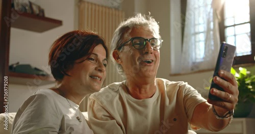 At home, a couple of grandparents make a video call to their relatives and friends using the smartphone . The elderly couple smiles, blows kisses and is happy to talk to them.