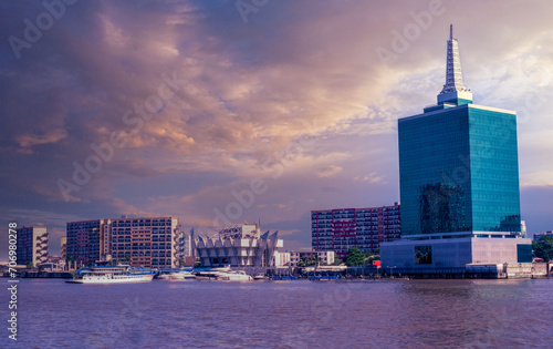 Golden Hour Cityscape with Modern Skyscrapers, Reflective Blue Building, and Boats on River