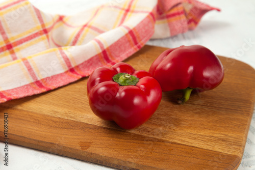 Cutting board with two whole red bell pepper and red kitchen towel on white wooden background. .