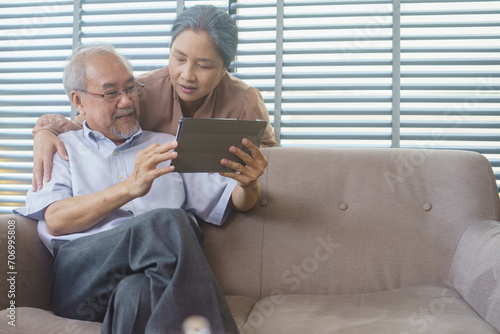 Asian grandfather and grandmother watching or meeting video call to family with digital tablet in the living room. Senior couple playing social media or searching on the internet.