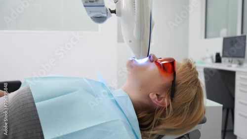 Close-up of female patient face at laser teeth whitening in the modern dental office. Blonde woman in protective red glasses bleaching teeth. Teeth whitening procedure with ultraviolet light UV lamp. photo